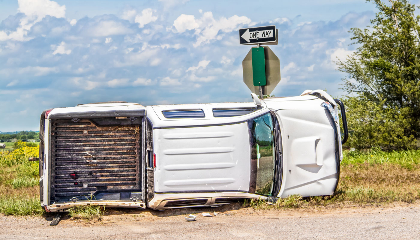 White long cab pick up truck turned on its side in a wreck at an intersection in the country beside a stop sign.