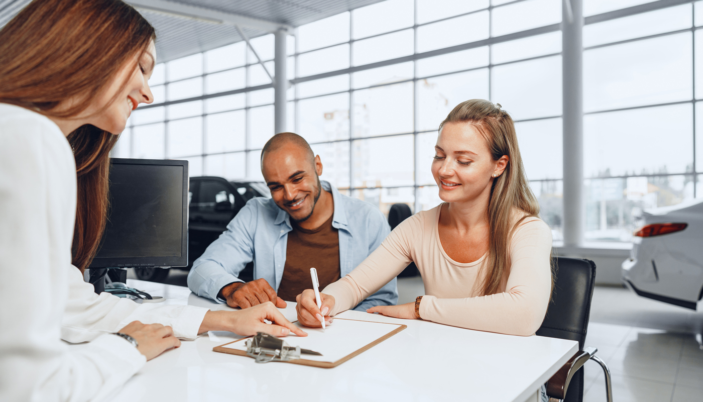 Beautiful young couple signs documents at car dealership showroom