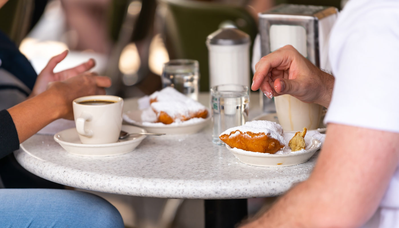 couple sitting outside at sidewalk cafe by table drinking chicory coffee and eating deep fried beignet donut powdered with sugar