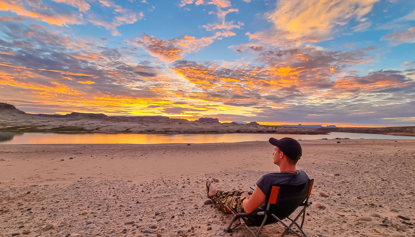 Man sitting on camping chair at Lone Rock beach campground with scenic sunrise view of Wahweap Bay at Lake Powell