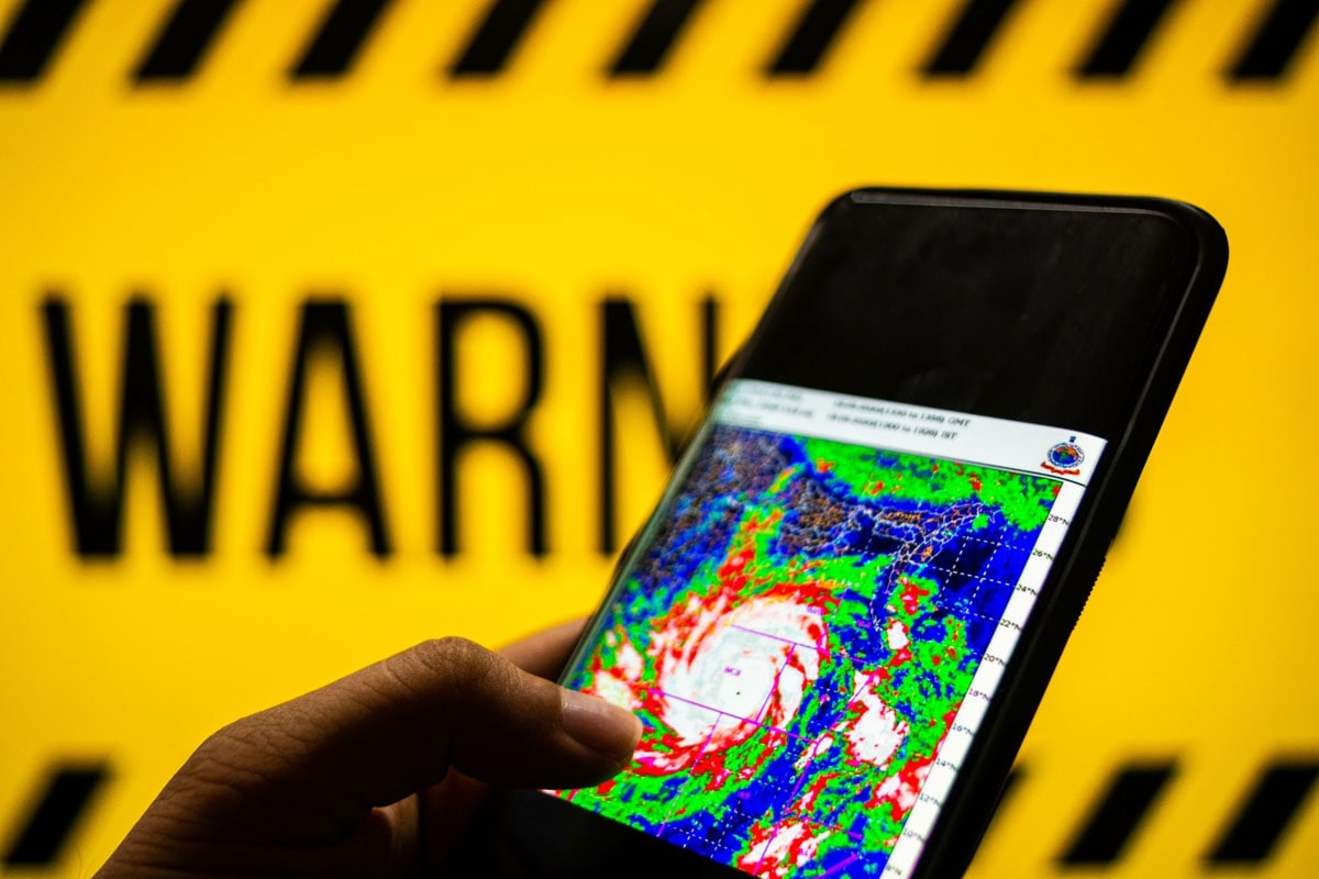 A phone in the foreground with the map of an approaching hurricane with a warning sign in the background.