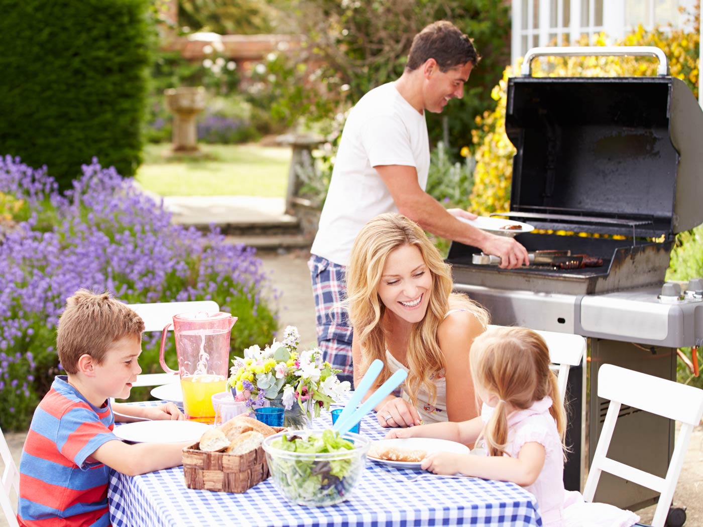 Family of four with dad grilling while mom and kids sit at table.
