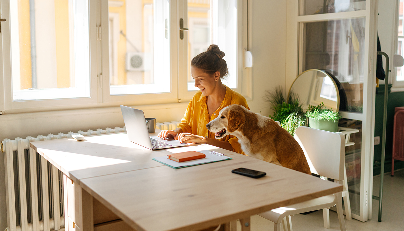 A woman smiles as she works at her laptop, with her dog seated next to her at the table.