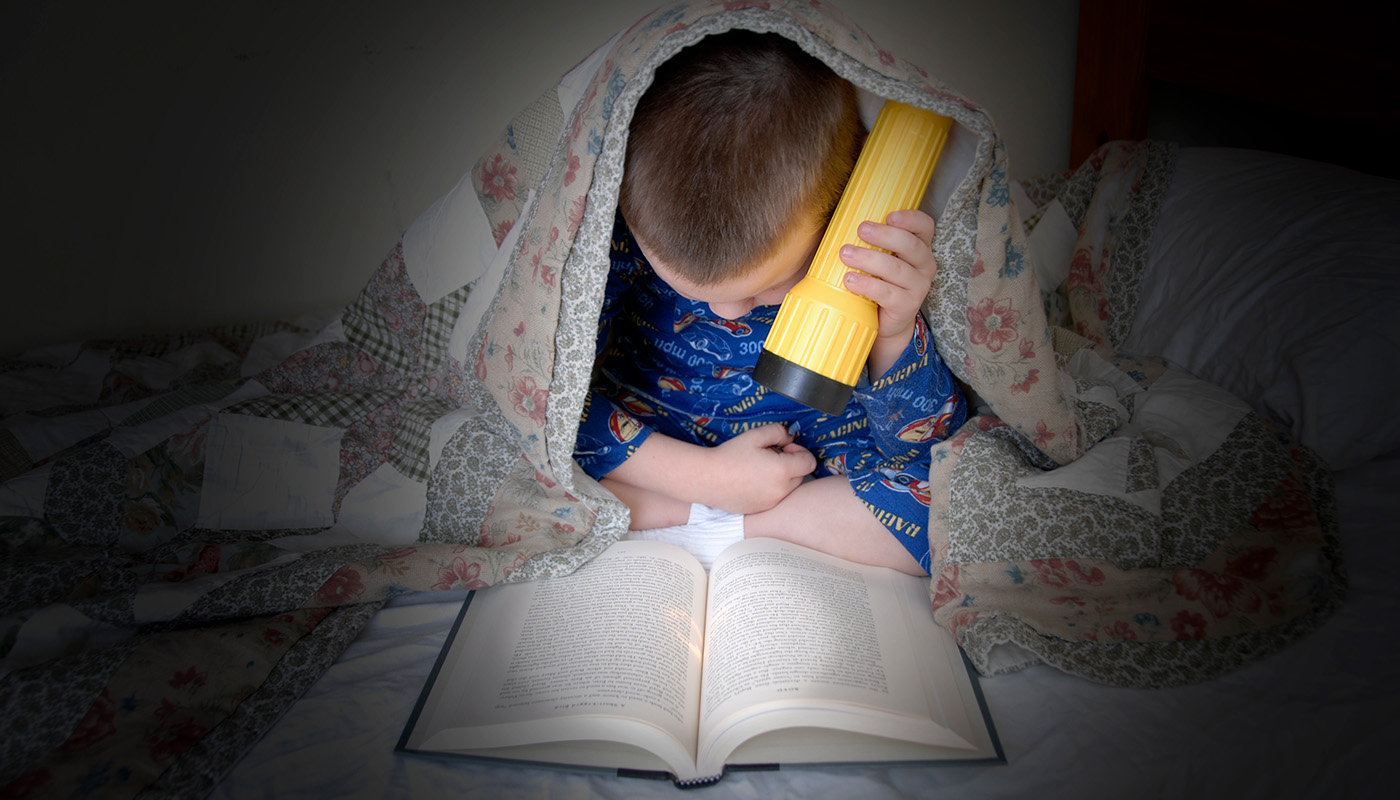 Young boy reading a book under covers with flashlight