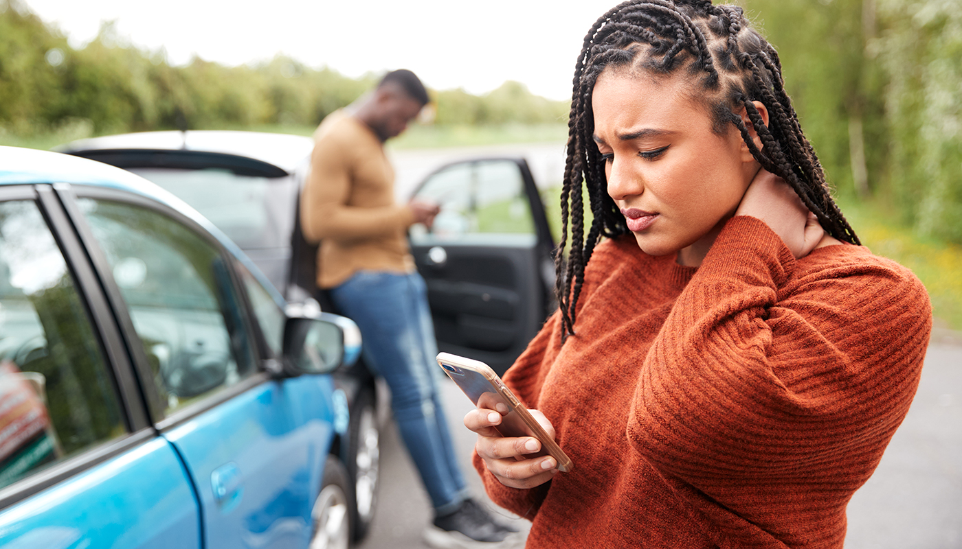 A woman with a worried expression looks at her smartphone with a car crash in the background.