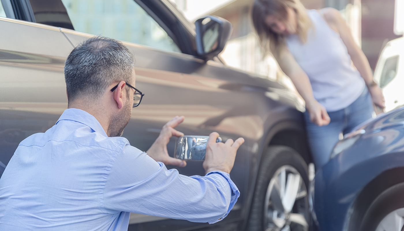 A woman points to crash damage on a car while a man takes pictures on a smartphone. 