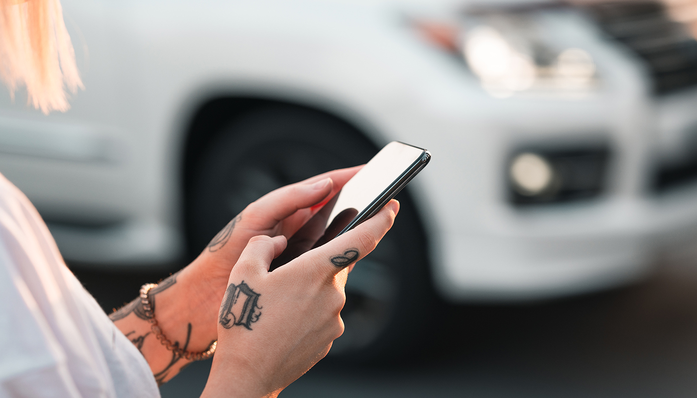 Someone with tattooed hands looks at their smartphone next to a vehicle. 