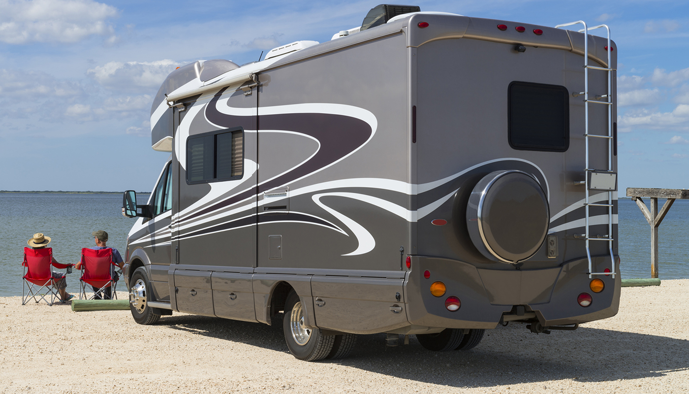 A couple sits on the beach looking out at the ocean. Their recreational vehicle is parked nearby. 