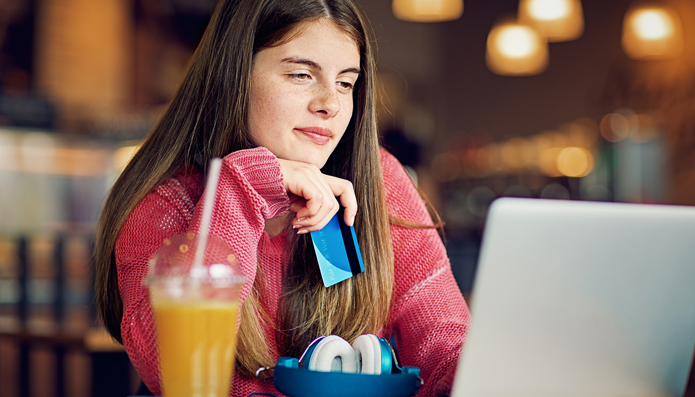 A teen girl holding a credit card while looking at her laptop in a cafe