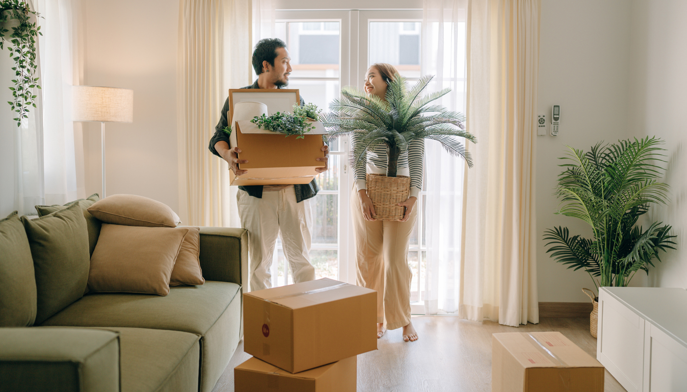 Couple moving boxes and plants into brightly lit house.
