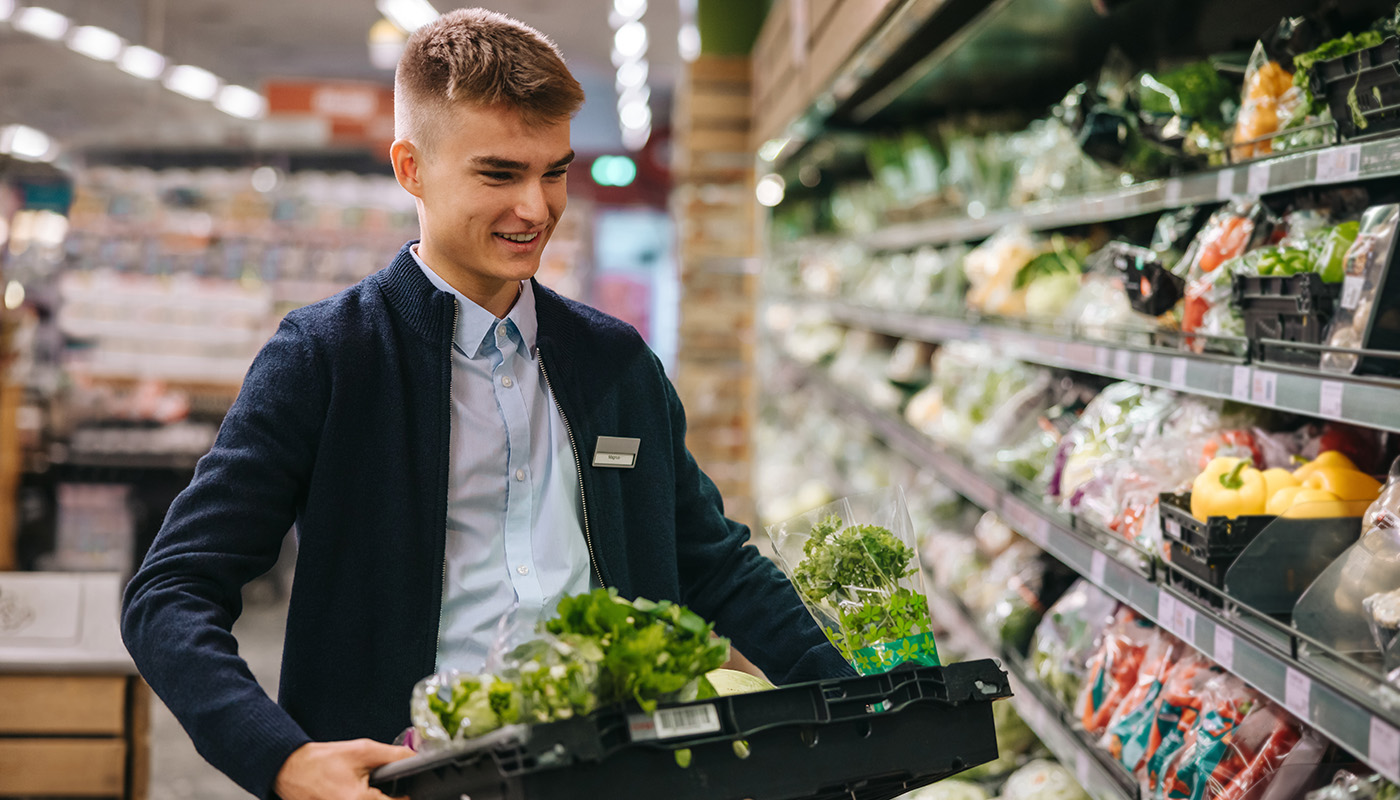 A young man loads fresh vegetables onto displays in produce section of grocery store