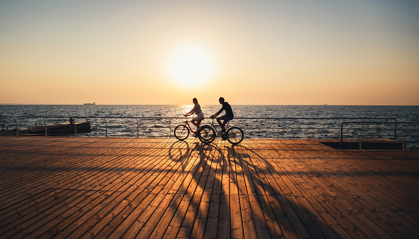 Two people riding bikes next on beach boardwalk with bright sun shining