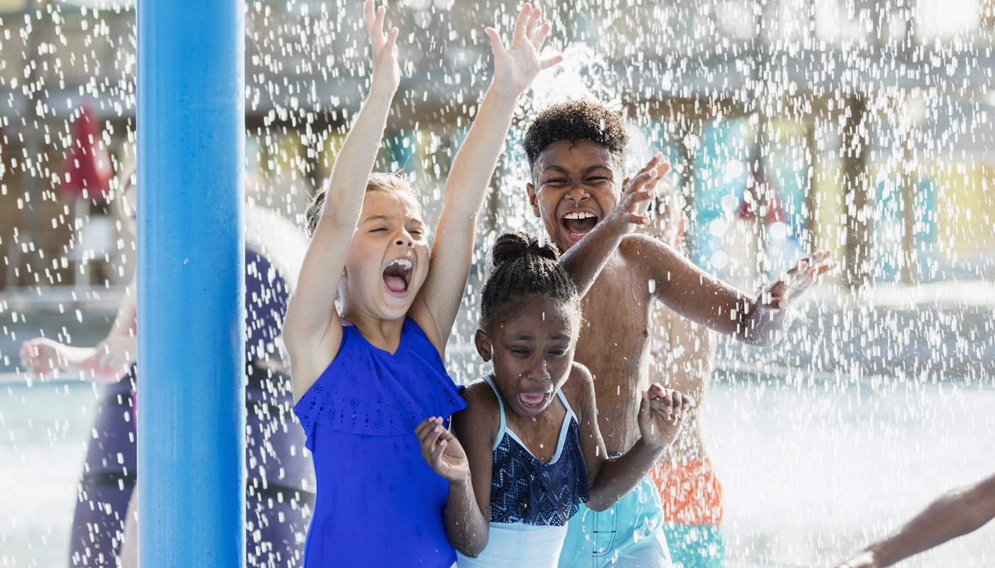 Multi-ethnic group of three children playing together at a water park. They are in a play area, standing under falling water