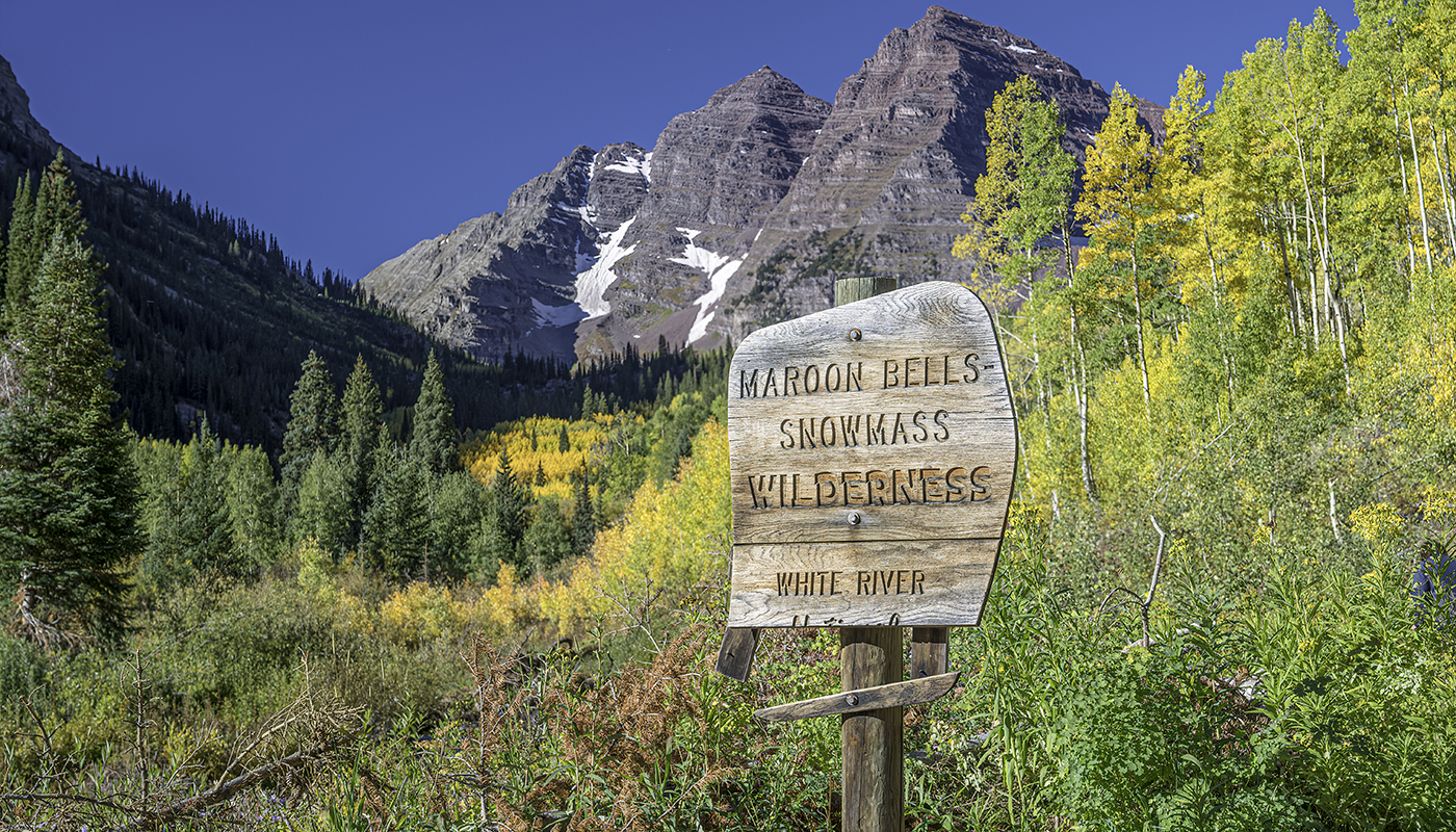 A wooden sign with mountains and foliage in the background reads, “Maroon Bells Snowmass Wilderness: White River.”