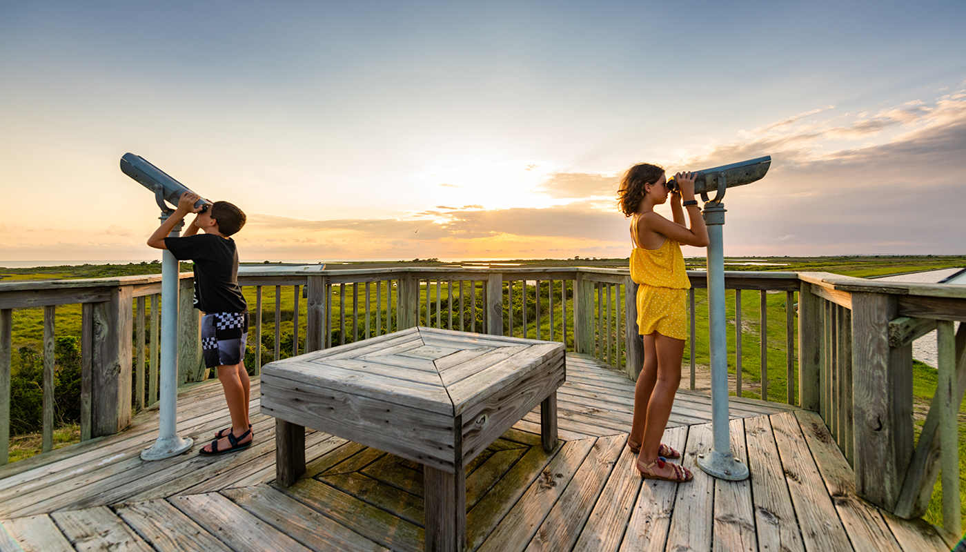  Two kids look through birdwatching telescopes on a wooden deck.