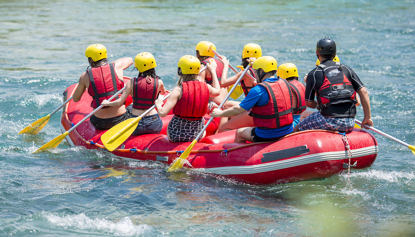 A group of nine people wearing life vests and helmets whitewater raft in a river.