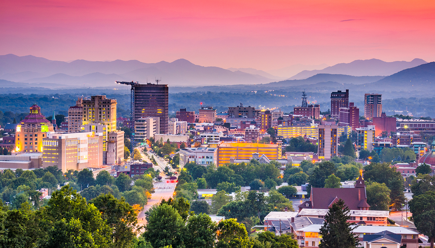 A lit up cityscape of Asheville with rolling hills and a colorful sunset in the background.