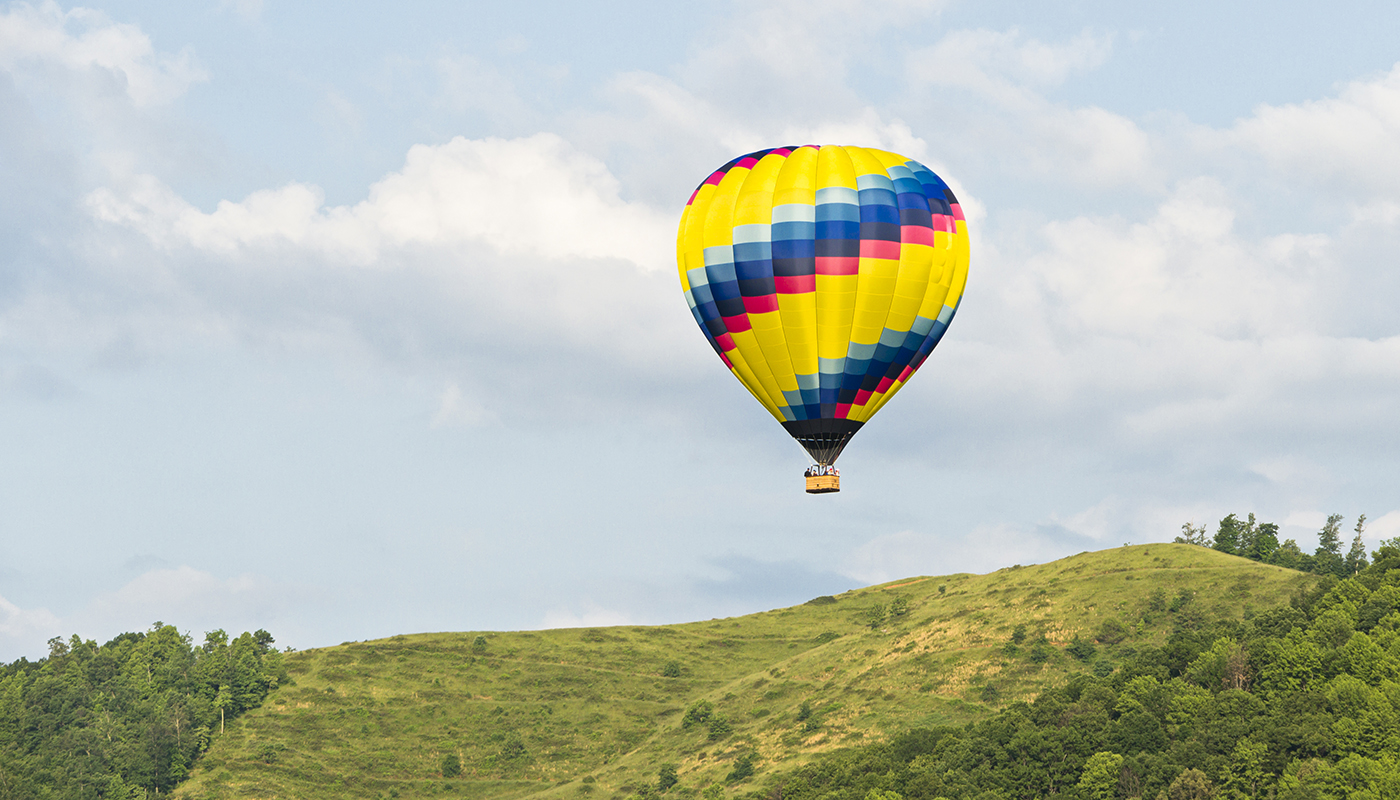 A yellow, blue, and red hot air balloon hovers over a hill with clouds in the background.