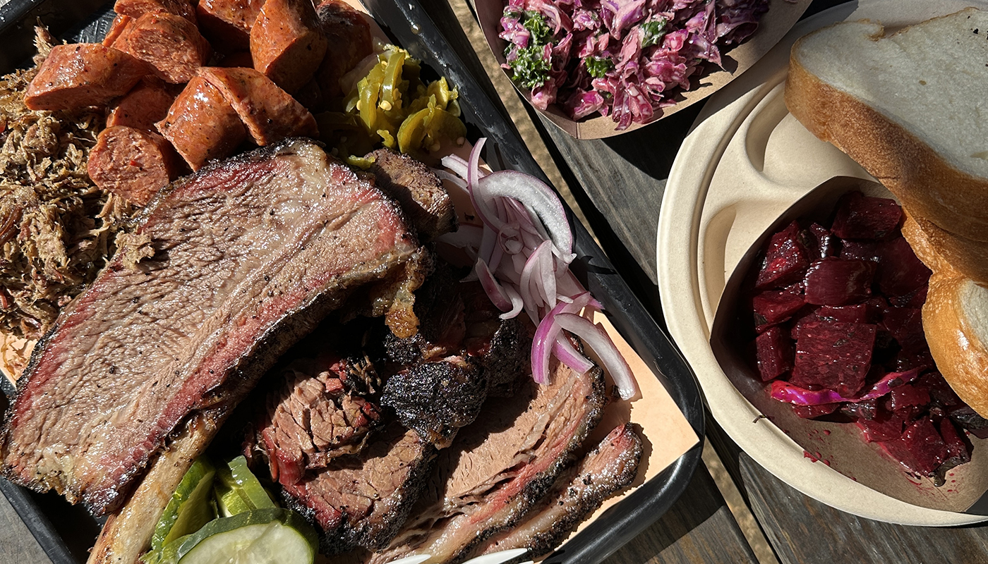 An overhead view of a large tray of Texas barbecue foods, including beef rib, beef brisket, sausage, pulled lamb, plus sides of pickles, mac and cheese, grits, beets, bread, and more.