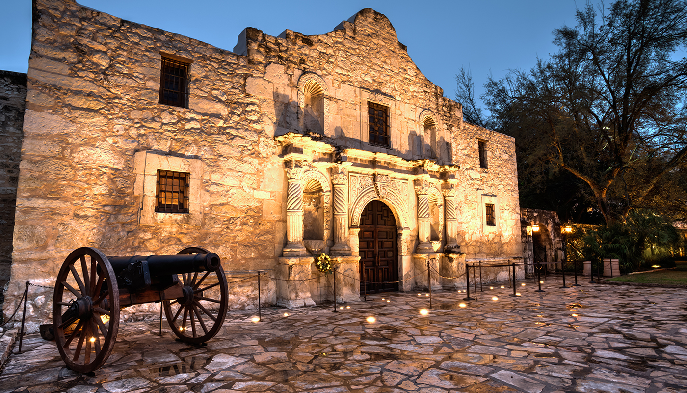 Cannon positioned in front of the Alamo, the façade is illuminated.