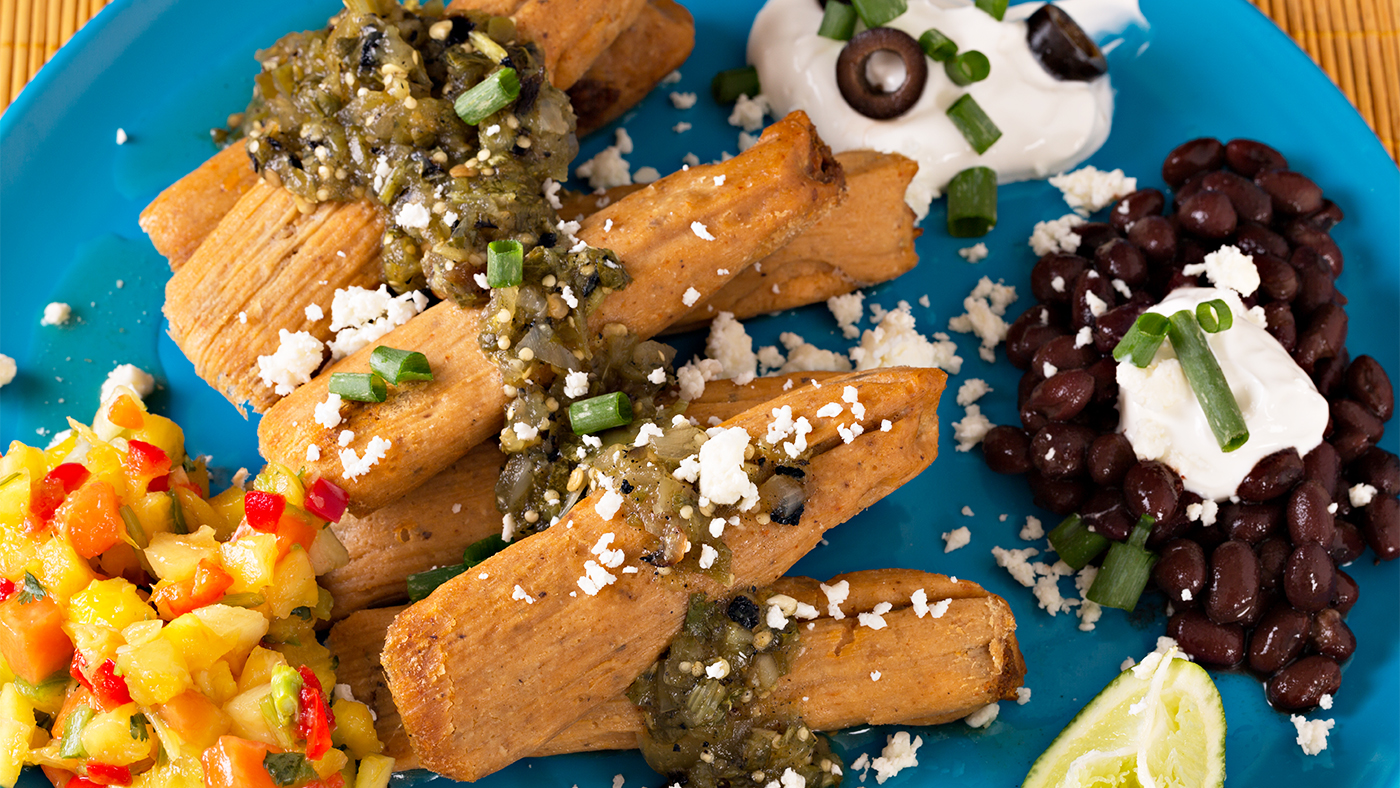 An overhead close up horizontal photograph of a blue plate choke full of pork tamales, black beans, tropical salsa, sour cream and garnished with chopped scallions, white cheese and black olives.