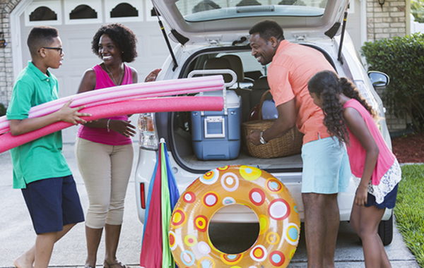 An African American family of four packing their car for a trip to the beach or pool, with cooler, umbrella and toys.  The teenage boy is carrying pool noodles and the 10 year old girl is watching the father put a picnic basket into the car.