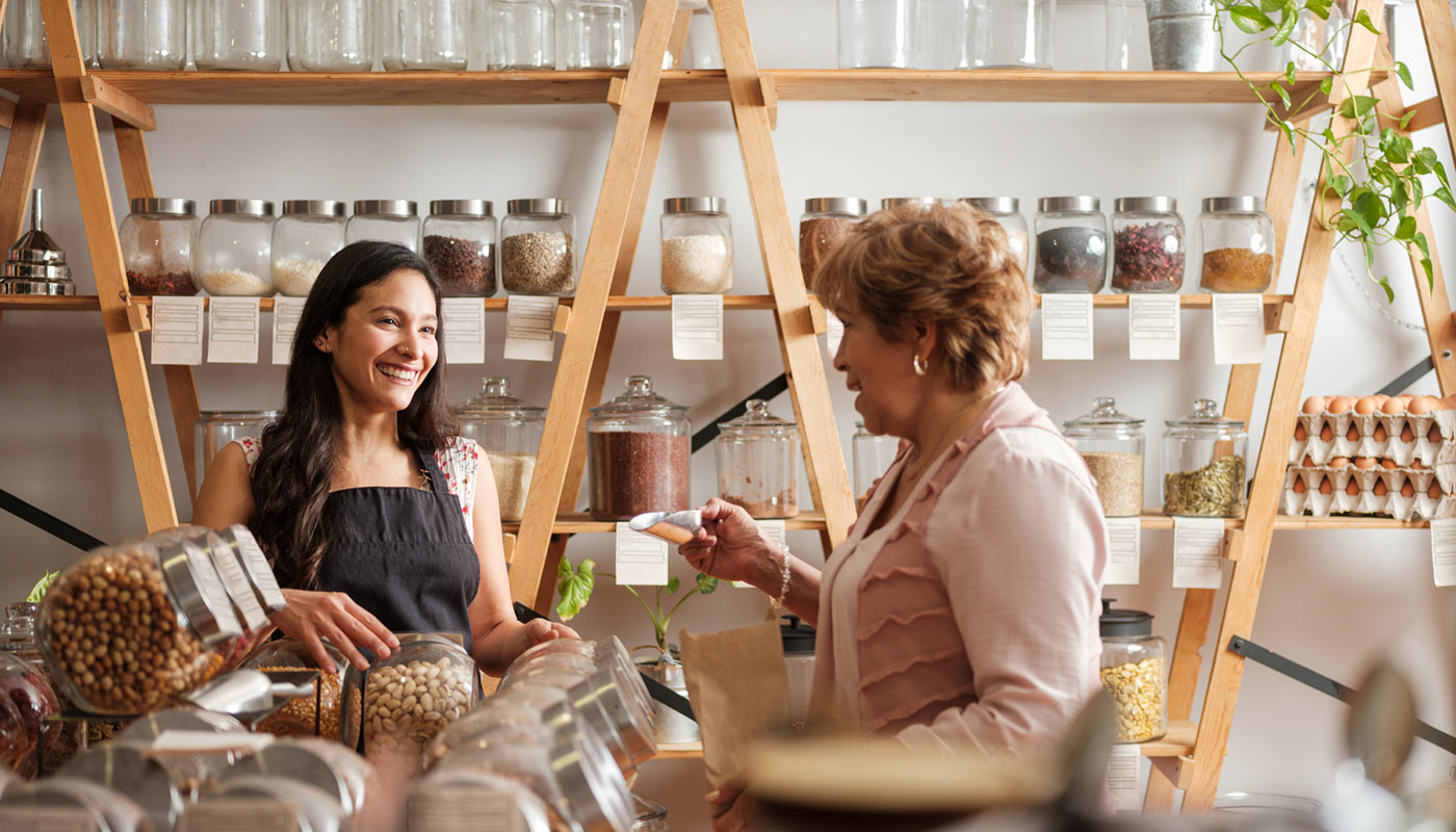 A woman scoops dry goods into a paper bag while conversing with a smiling woman.