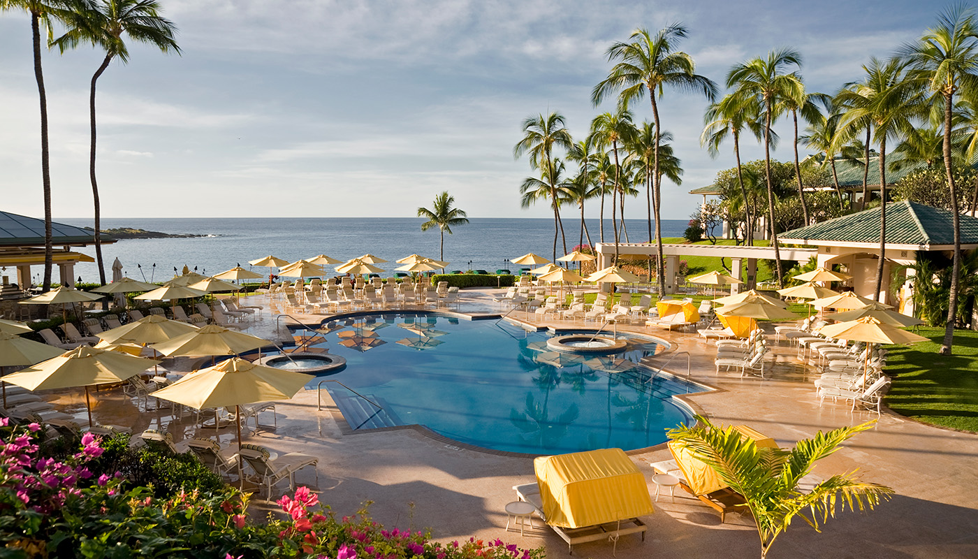 View of the ocean from the pool area of a resort in Hawaii. The pool is surrounded by lounge chairs, umbrellas and palm trees