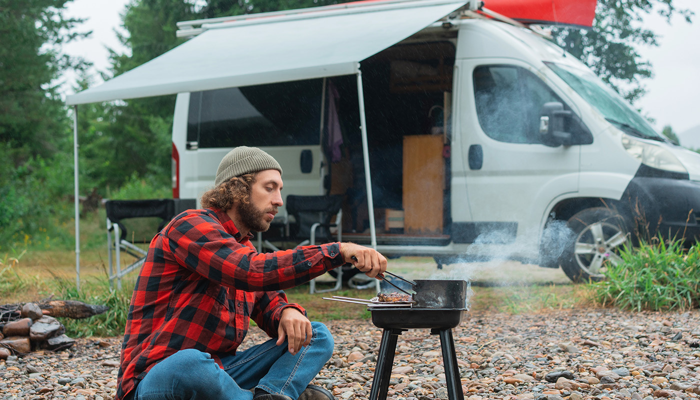 A man grills food on a small outdoor stove with his van in the background. 