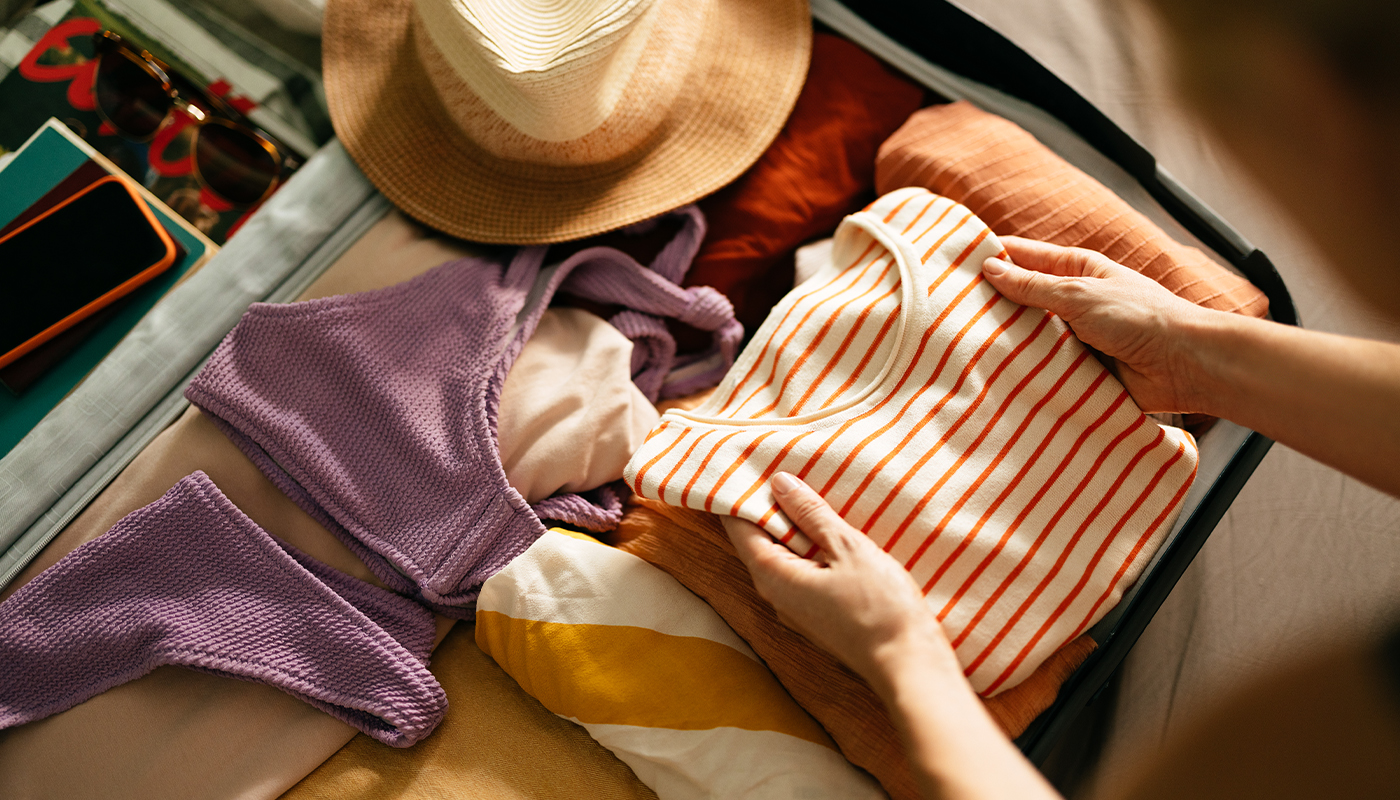 A pair of hands packs a shirt, swimsuit, hat and more into a suitcase. 