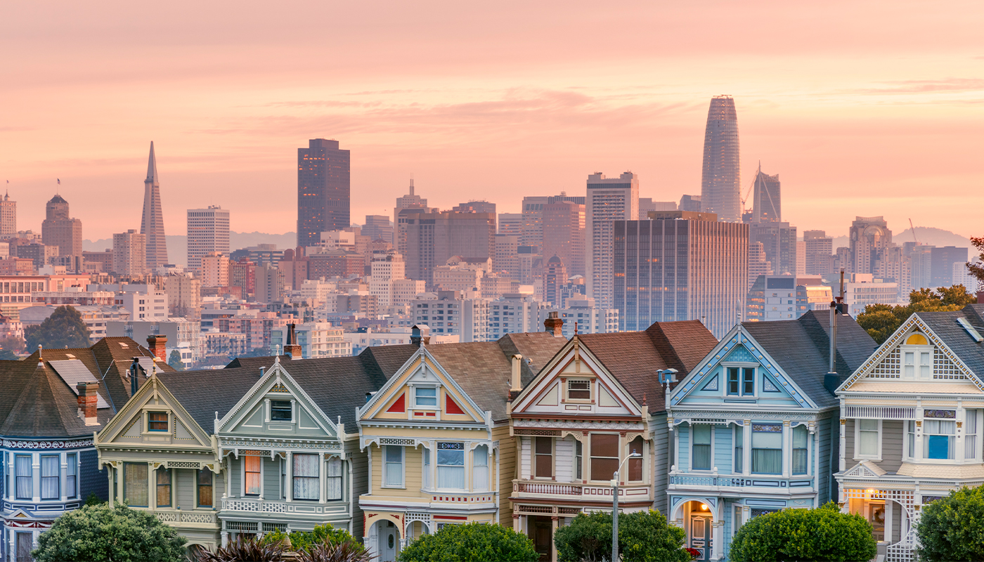 A historic neighborhood full of colorful houses packed tightly together in a row with San Francisco skyscrapers in the background.