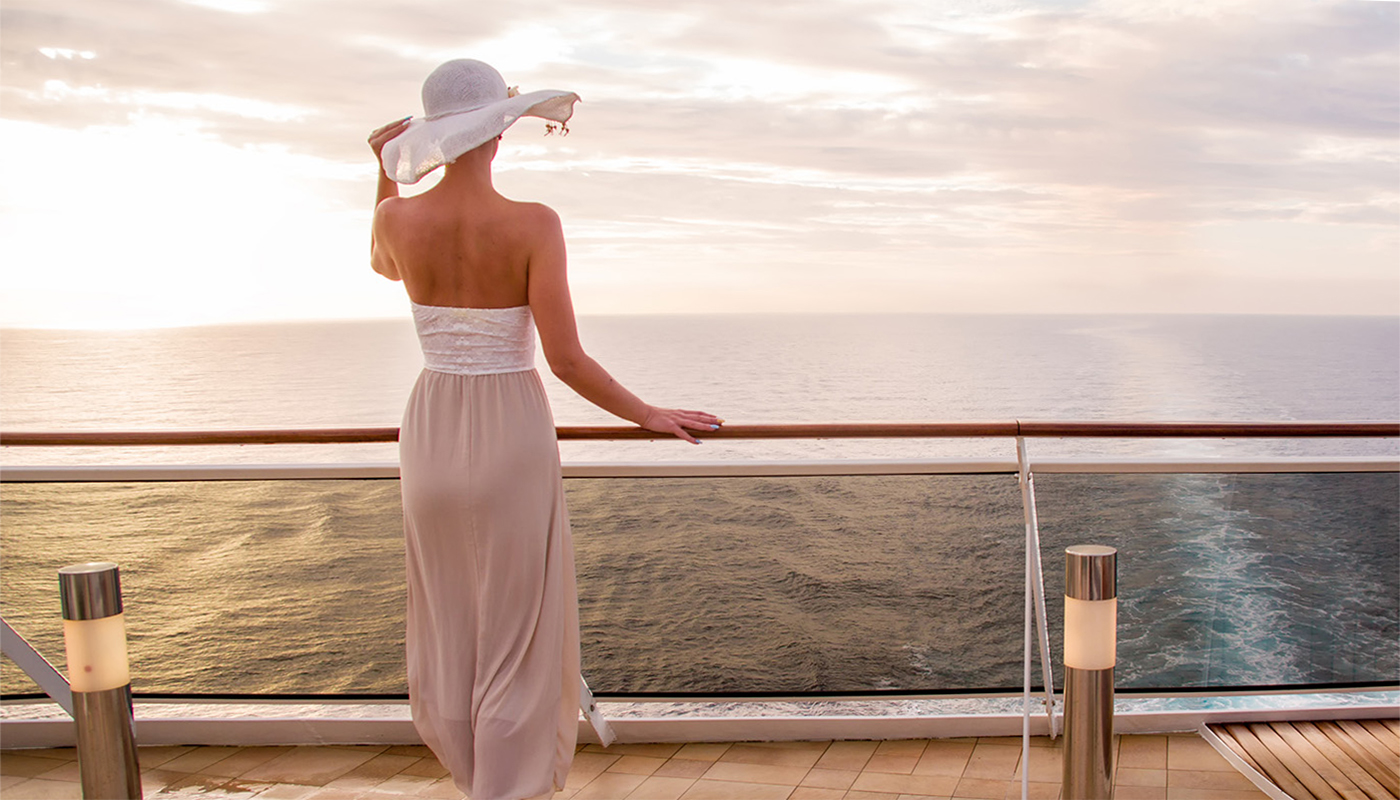 A woman in stylish cruise wear stands at a ship's railing and looks out at the ocean. 
