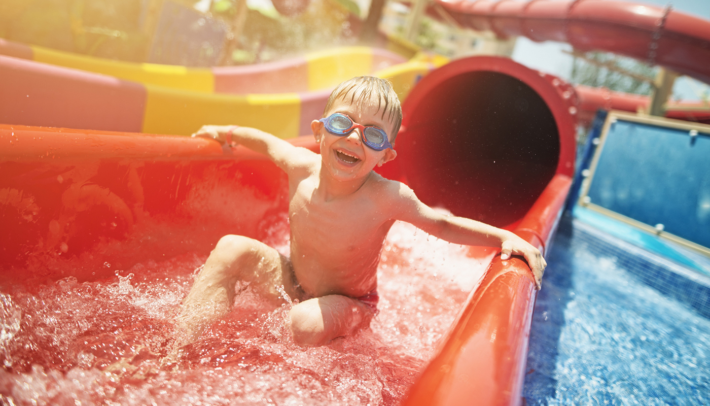 A young boy wearing swim goggles smiles as he emerges from a water slide