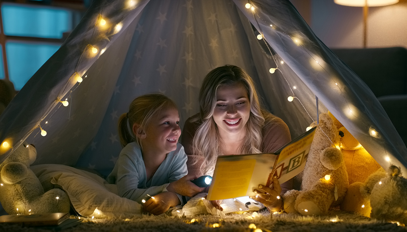 A mom and daughter read a book together as they lie inside a homemade tent