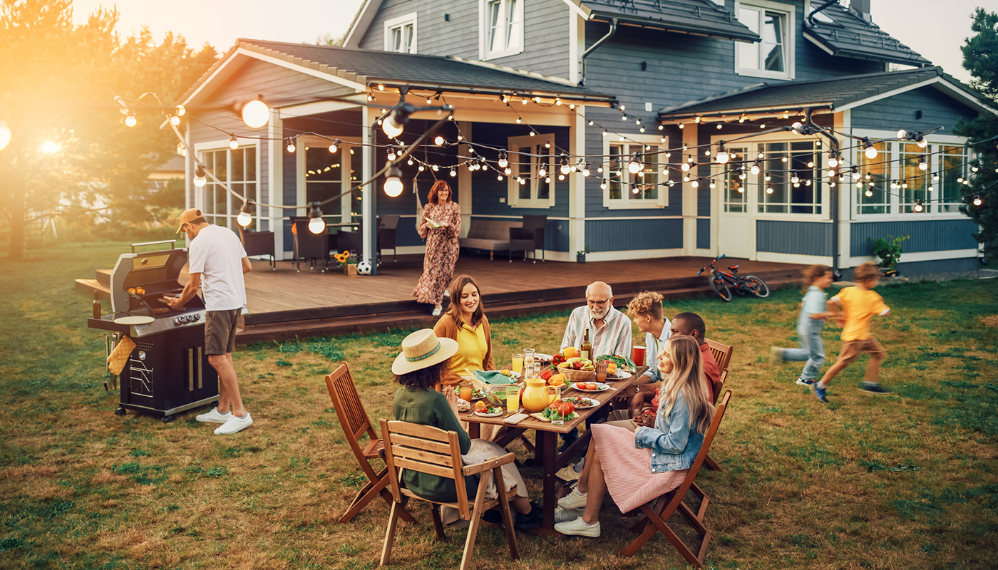 A family enjoys a meal at a table in the backyard while kids play and an adult grills food