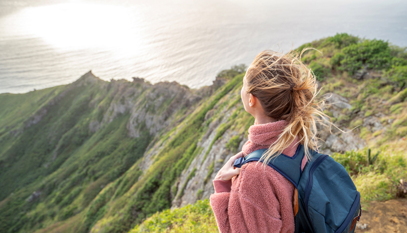 A hiker with a backpack looks out across the grass-capped hills of Hawaii. 