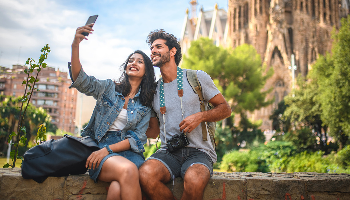 Male and female tourists sitting on wall in Barcelona taking a selfie