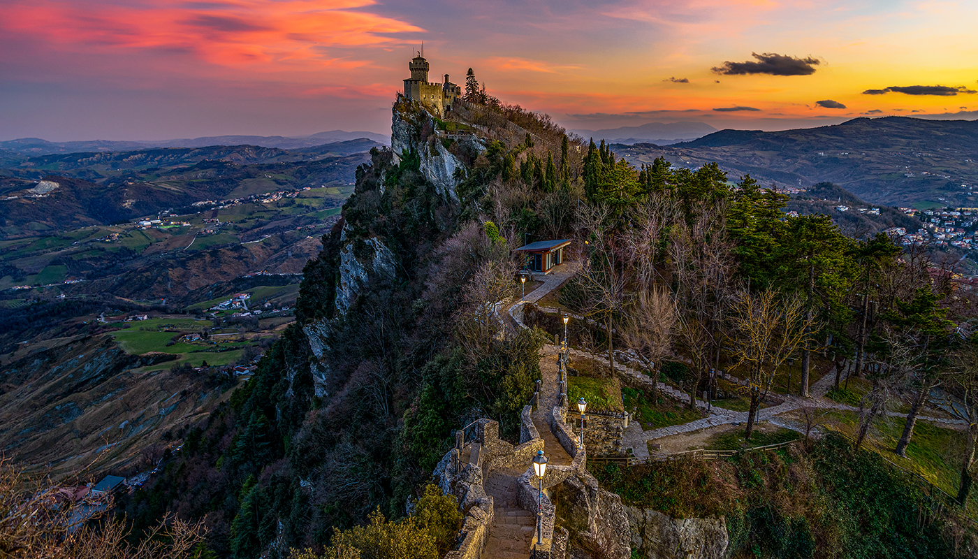 In San Marino there are 3 towers on the mountain ridge in a row. This is Falesia the second tower. The image was taken at sunset. The sky is filled with colorful clouds and the lanterns in the foreground have already been lit. The Tower tits on a peak. The terrain left to the tower drops steep.