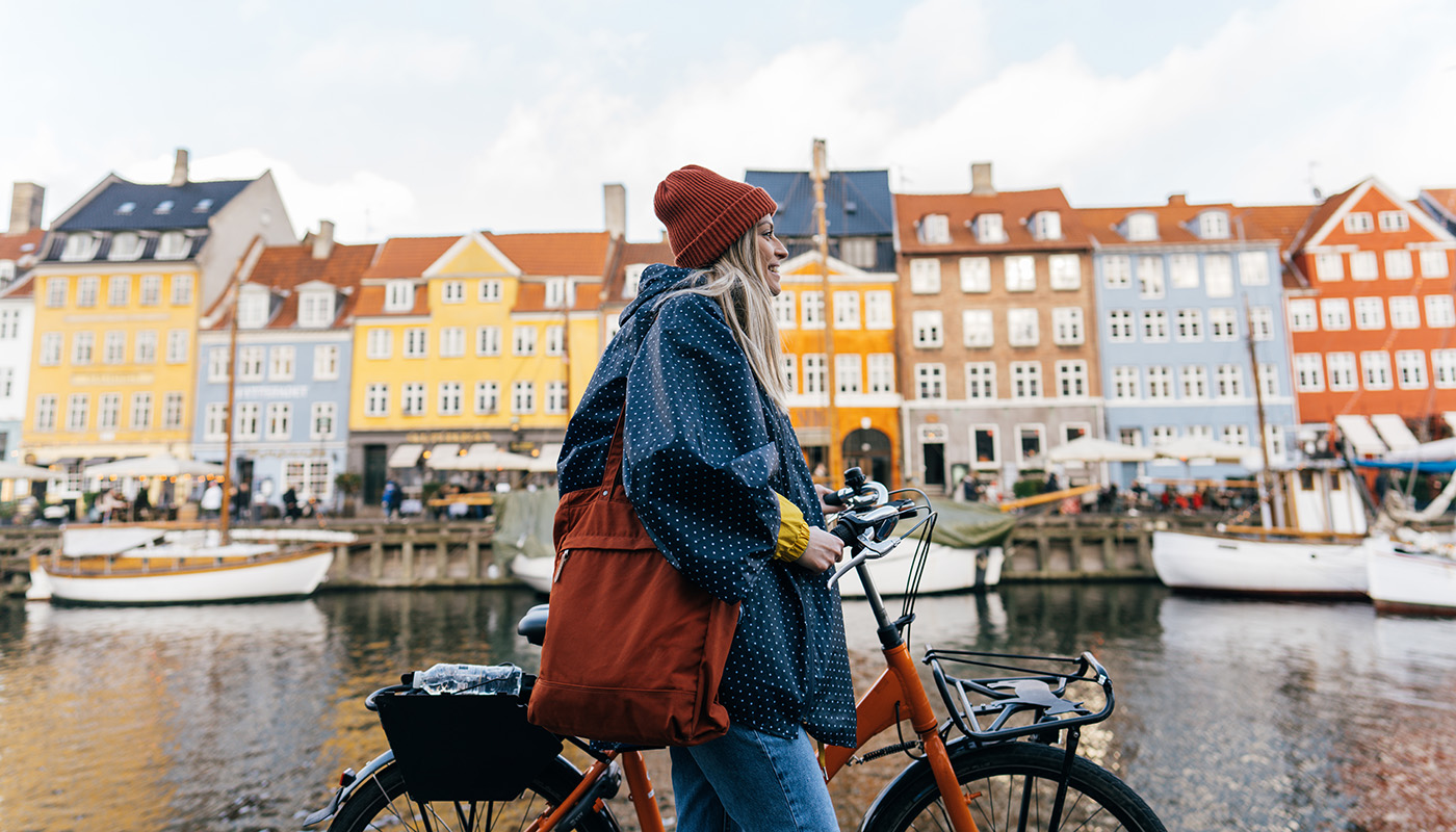 Young woman in hat with bike walks by colorful homes along water in Copenhagen