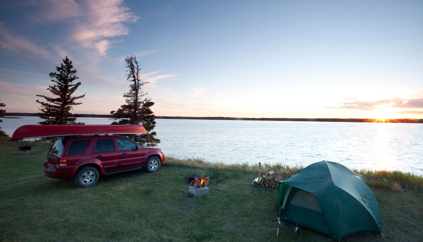 Red SUV with canoe on top parked by a tent and a large open lake.