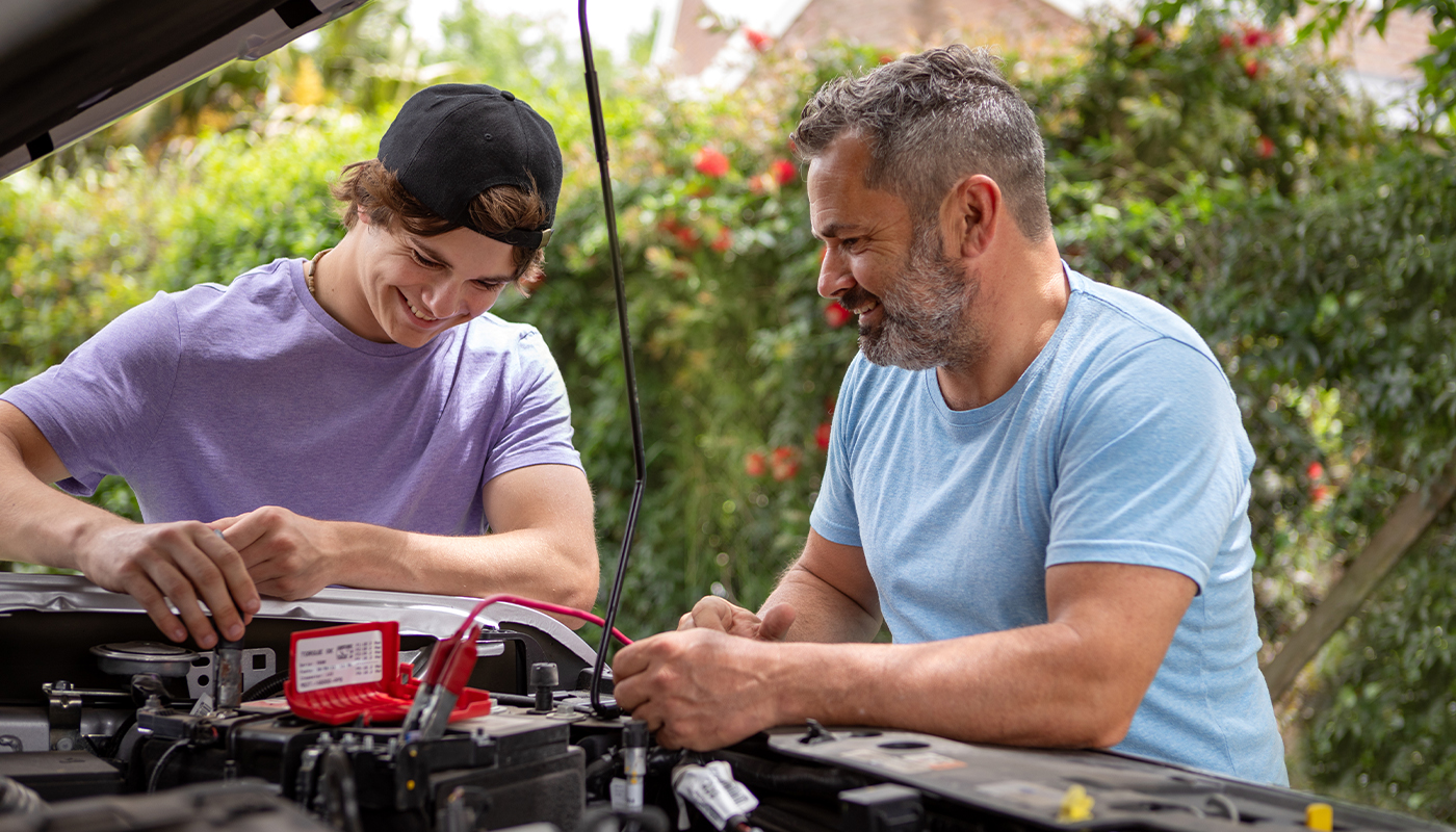 Two men check a battery under the hood of a vehicle.