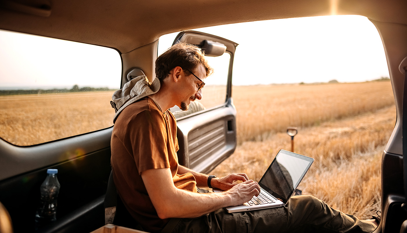 Young man sitting in his vehicle with the door open, typing on his laptop next to a field