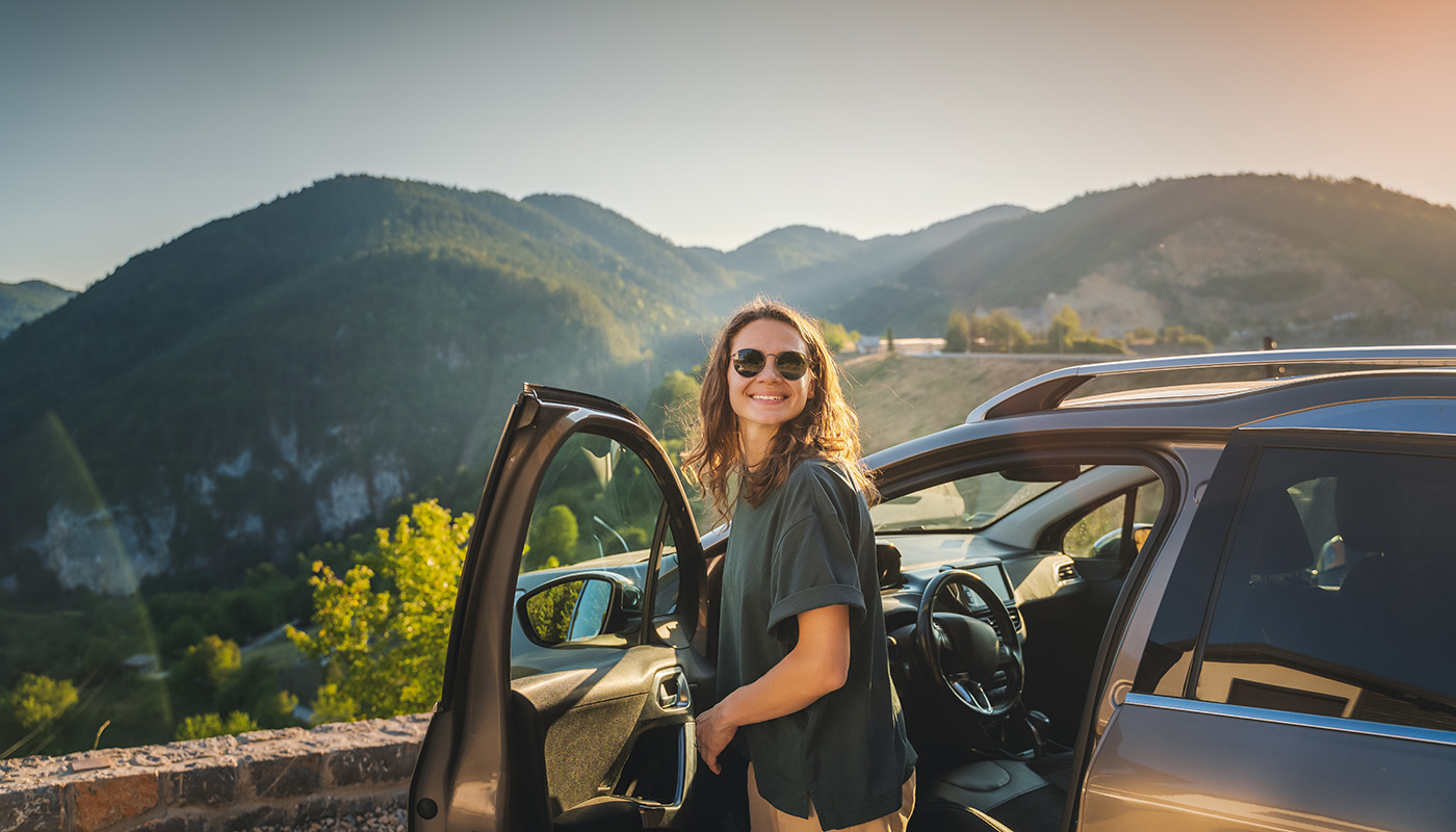 Smiling young woman getting out of car with mountains in the background