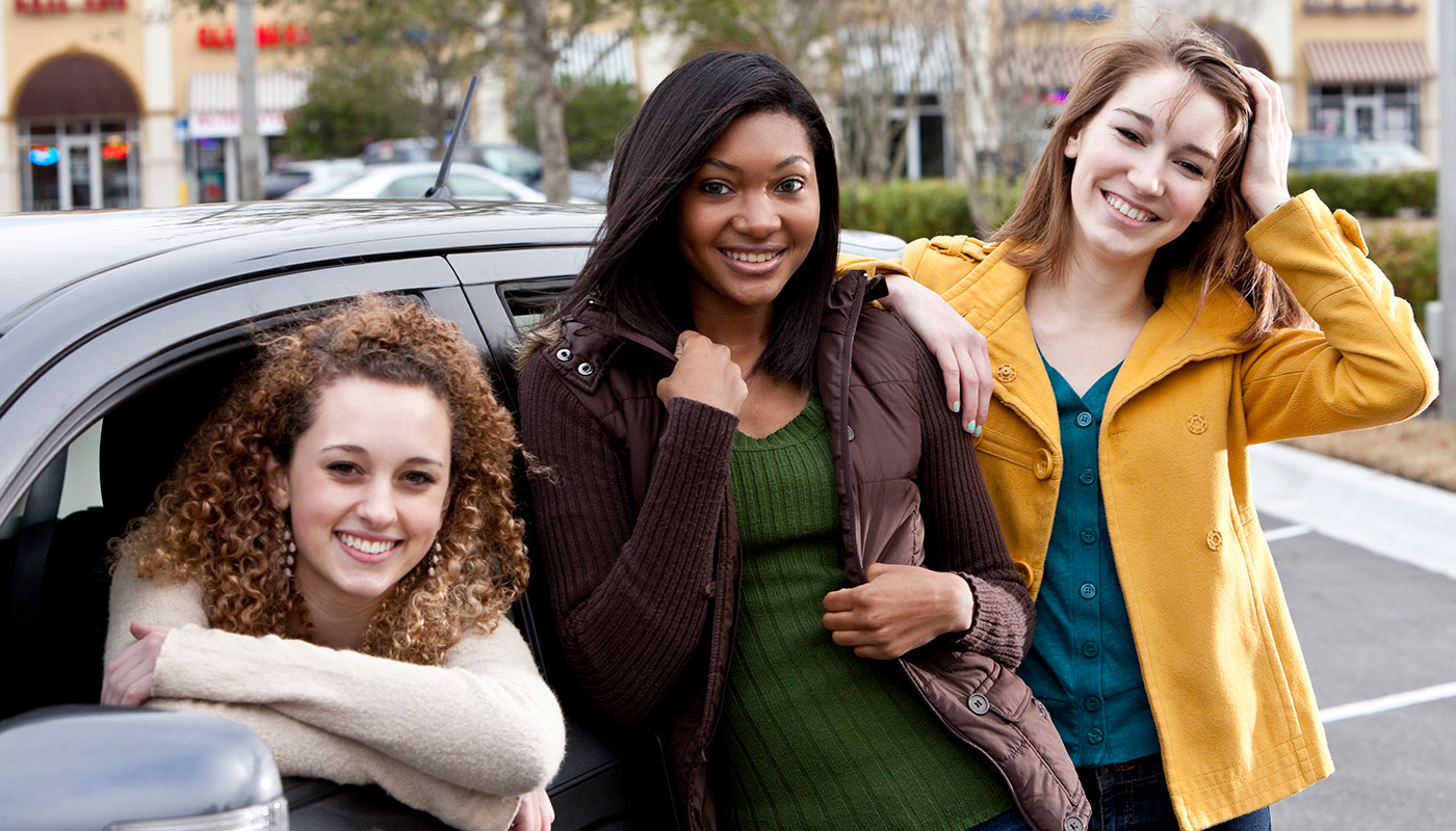 Three teenagers smile in front of a vehicle.