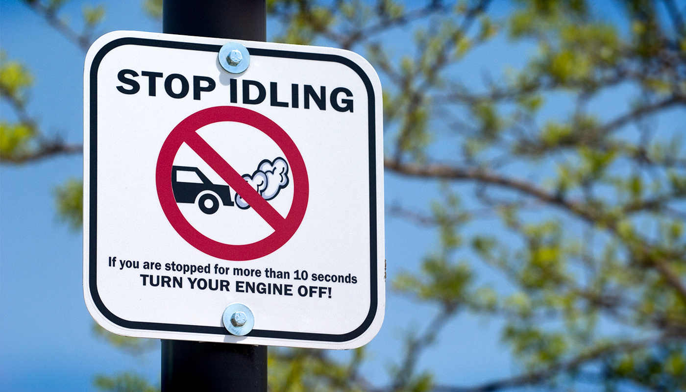 A white sign reads, “STOP IDLING.” It has a crossed-out image of a car emitting fumes into the air. At the bottom of the sign it reads, “If you are stopped for more than 10 seconds TURN YOUR ENGINE OFF!”