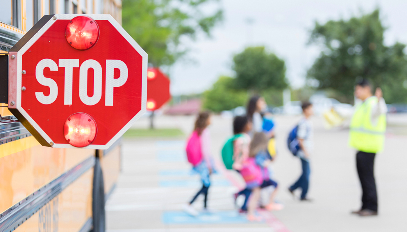 Focus is on a flashing school bus stop sign in the foreground as a group of schoolchildren cross a parking lot with the help of a crossing guard in the distance.