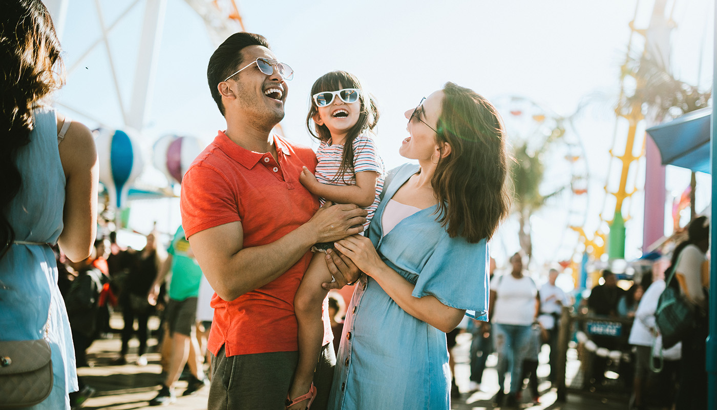 Family smiling outside on a sunny day with rides and activities in the background