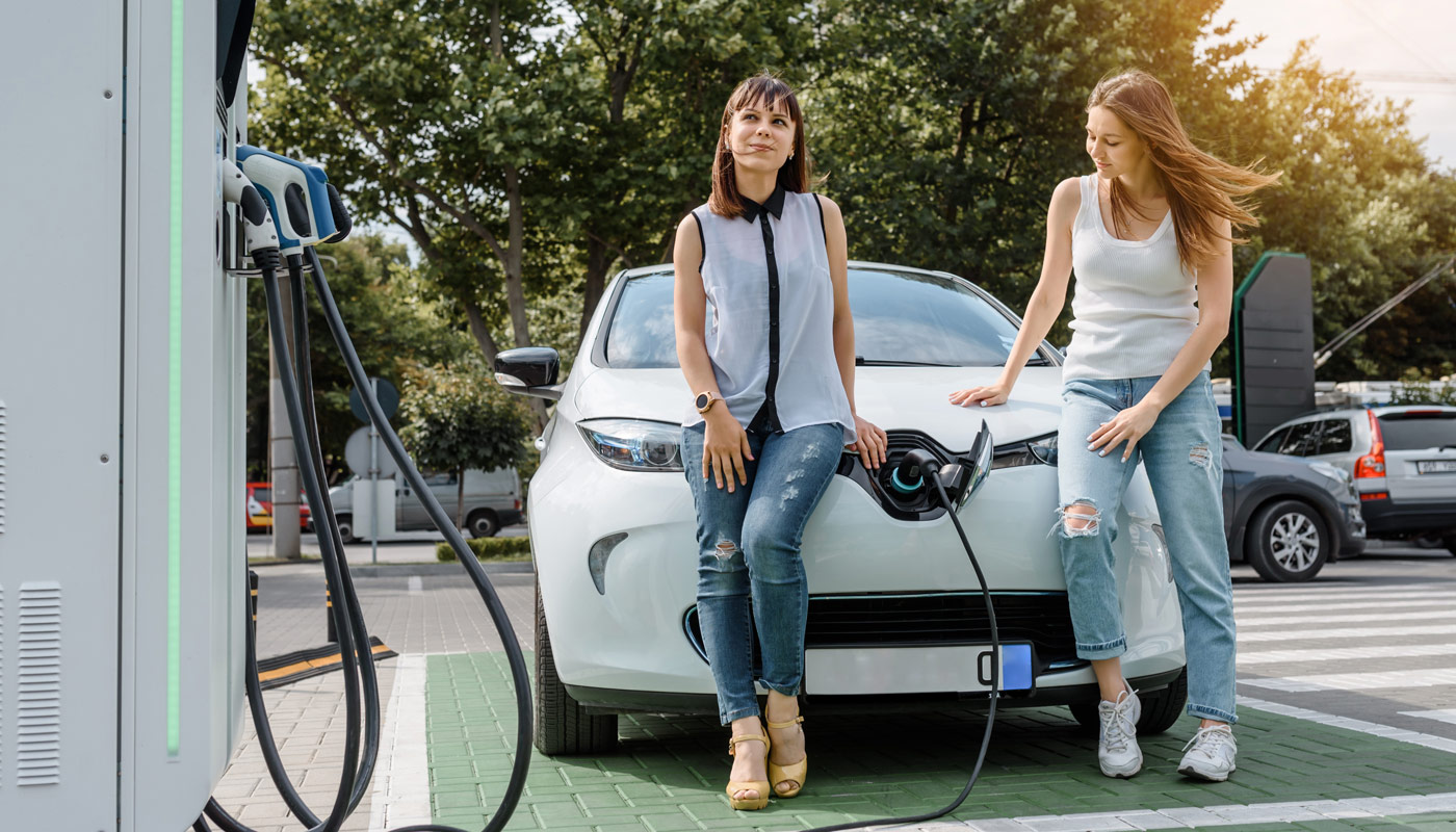 Two happy young beautiful women are talking to each other until their electric car is charging at the charging station situated in the car park.