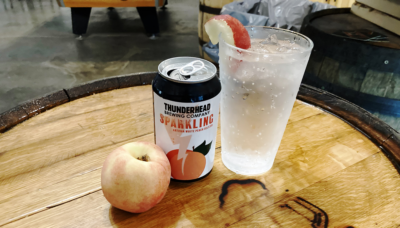 Open can of Thunderhead Brewing Company Sparkling Peach Seltzer next to a filled glass with peach garnish and a whole peach