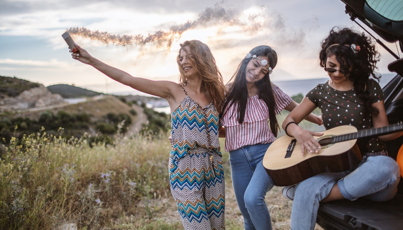 3 Hippie girls behind a vehicle with guitar.